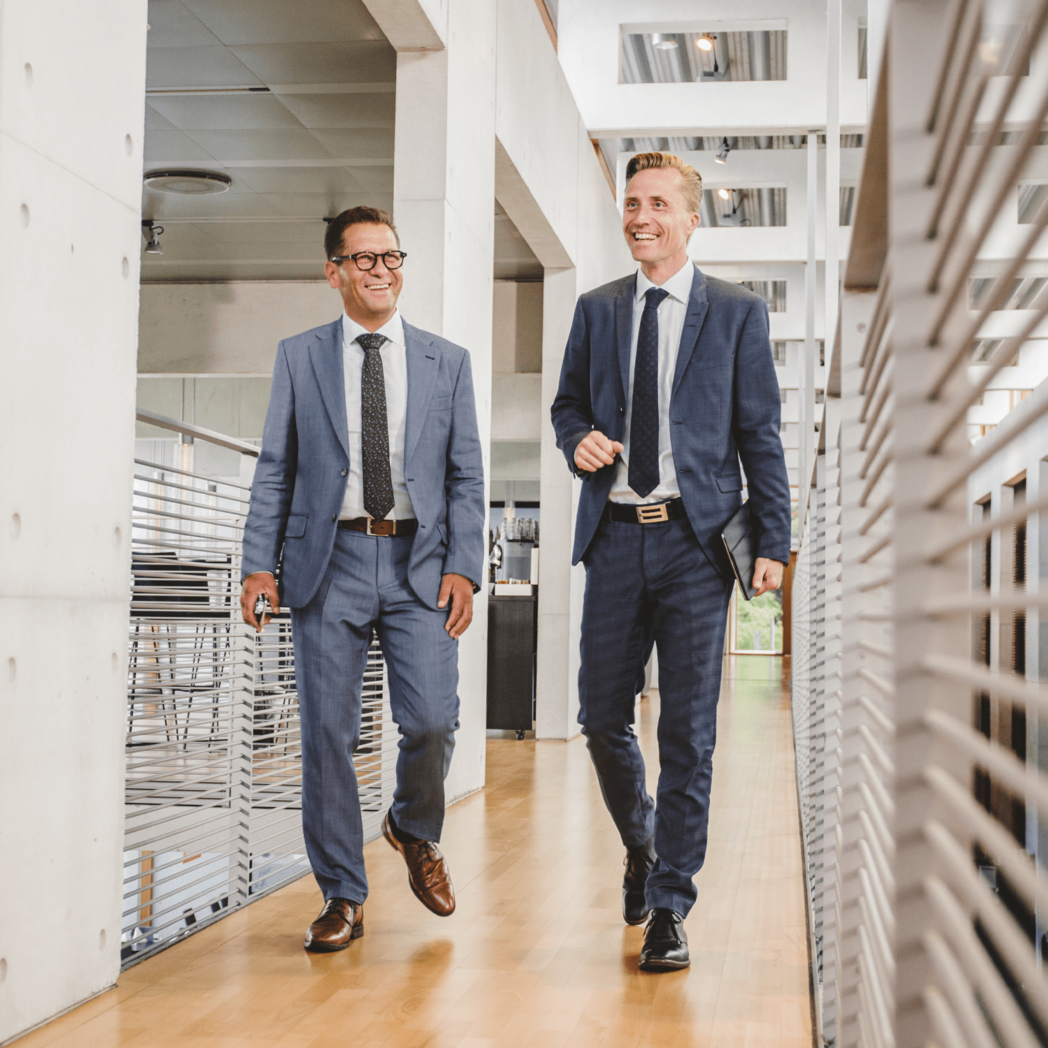 Two professionally dressed men in business suits walking and smiling in a modern office building, representing a positive and professional work environment.