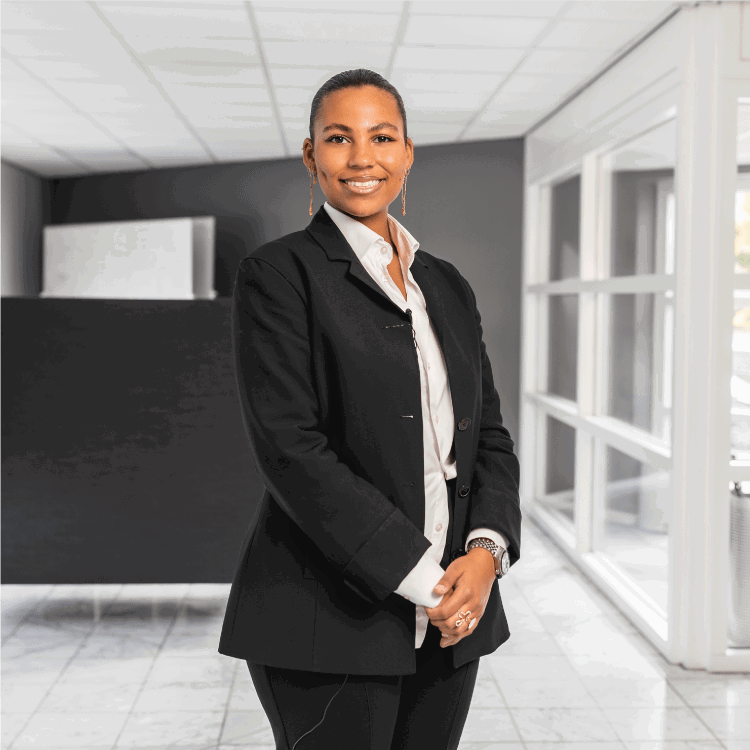 A confident professional woman in a black blazer and white shirt stands in a modern office space, smiling warmly. The background features a sleek reception area with large windows and a tiled floor.