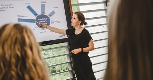 Presenting Data: A woman in black attire standing in front of a large screen, pointing at a presentation slide displaying a circular data visualization, while an audience listens attentively.
