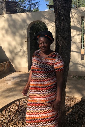 A woman wearing a patterned dress and a black headwrap stands under a tree, with a beige arched structure and shadows in the background. She has a calm and content expression.