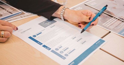 Showing Results: A person’s hand holding a pen and pointing at a document with structured information on a table, surrounded by other documents and brochures.