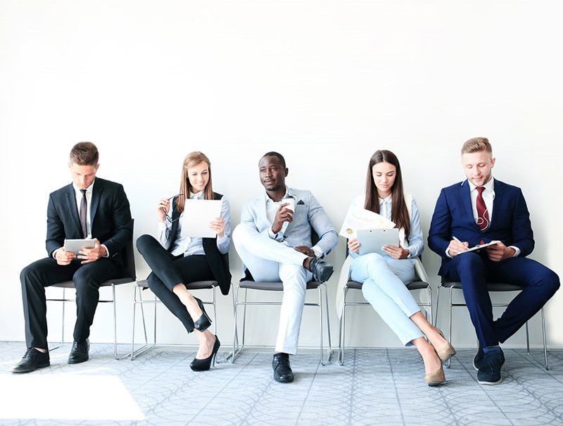 Five professionally dressed individuals are sitting in a row on chairs, appearing to be in a waiting area. They are reviewing documents, using tablets, or waiting, suggesting a job interview or business setting.