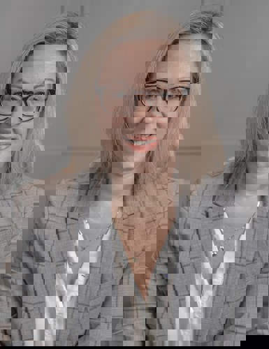 A professional headshot of a smiling woman with shoulder-length blonde hair and glasses, wearing a gray plaid blazer over a white blouse, against a neutral background.