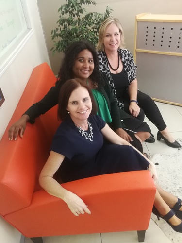Three women sitting together on an orange couch in an office lounge area. They are dressed in professional attire and smiling at the camera, with a modern reception area in the background.