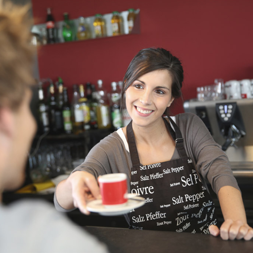 A smiling barista wearing a black apron serves a small red cup of coffee to a customer in a café with a well-stocked bar in the background.