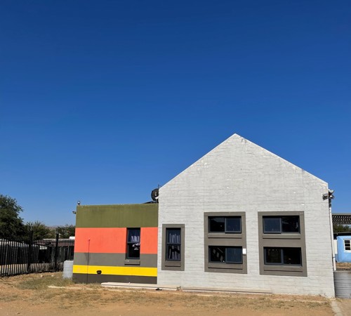A completed building with a white gabled roof and modern black-framed windows stands beside a smaller structure painted in bright horizontal stripes of red, orange, and yellow. The setting is in a dry, open area under a deep blue sky.