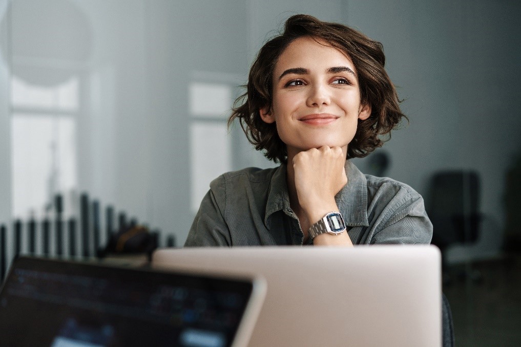A young woman with short brown hair and a smile sits at a desk in a modern office, resting her chin on her hand while looking thoughtfully. A laptop is open in front of her, and the background features a glass-walled meeting room with minimalist decor.
