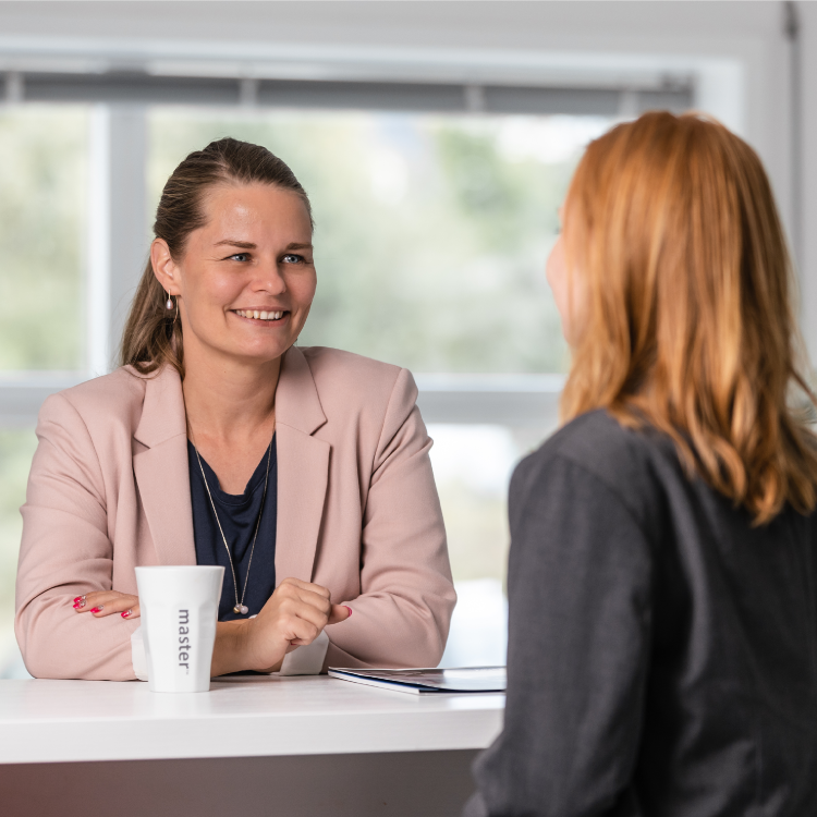 Two professional women engaged in a conversation at a modern office. One woman, wearing a pink blazer, smiles while sitting at a white table with a branded 'Master' coffee mug and a notebook.